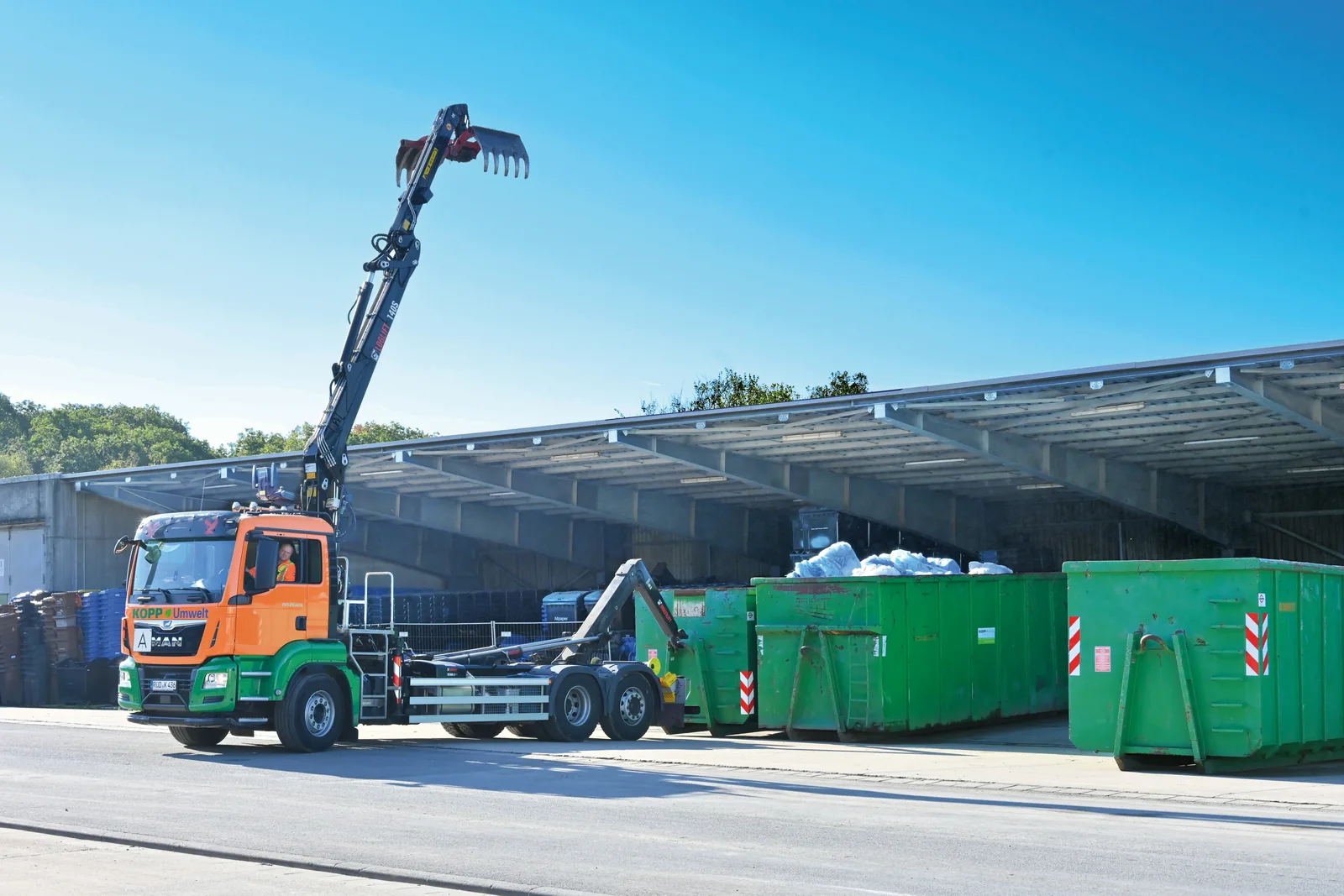 A hook-lift truck beside green roll-off containers at a depot